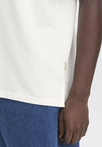 Camiseta de algodón blanca con una abertura lateral, que presenta una pequeña etiqueta en la parte inferior izquierda. Combinada con pantalones de mezclilla azules. La textura parece suave.