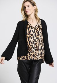 Schwarze gestrickte Strickjacke mit weiten Ärmeln, getragen über einer Bluse mit Leopardenmuster. Das Model trägt schwarze Lederhosen vor einem schlichten Hintergrund.