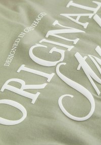 Pano bližnji posnetek svetlo zelene tkanine z dvignjenim belim napisom "ORIGINALS" in manjšim napisom "OBLIKOVANO V KOPENHAGENU."