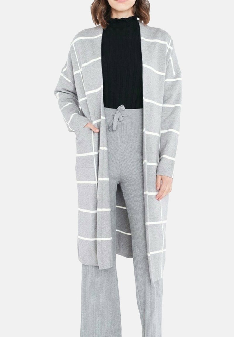 Cardigan gris avec des rayures blanches, à manches longues, ouvert à l'avant et avec des poches latérales, superposé à un haut en maille noir et un pantalon en tricot gris.