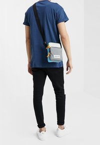 Grå crossover-taske med geometrisk design, justerbar rem og orange detaljer, båret over en blå skjorte og sorte bukser.