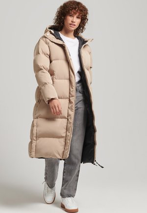 Woman wearing long beige puffer coat over white shirt and gray jeans, walking forward against neutral background.