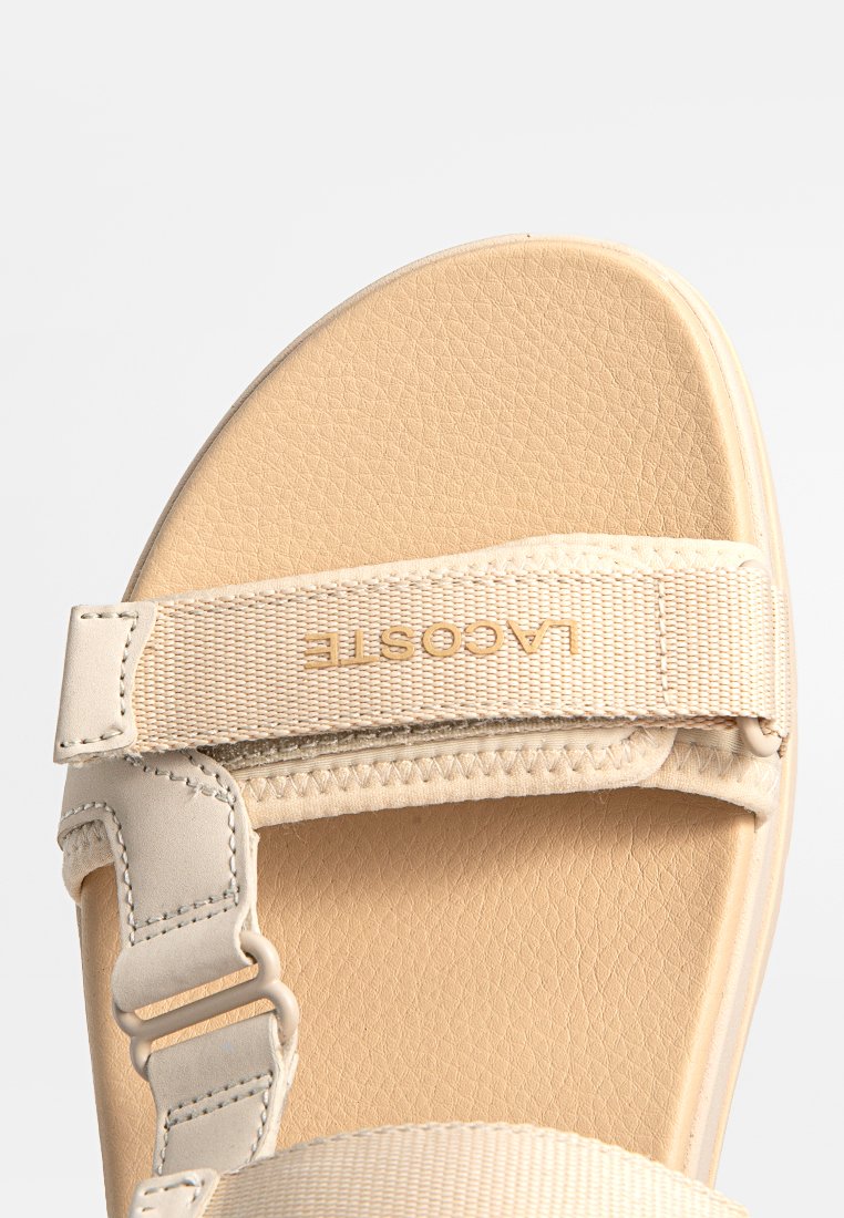 Sandale beige avec semelle intérieure texturée, brides de soutien et détail de logo. Les brides sont ajustables et fabriquées en un matériau en tissu durable.