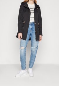 Svart huva parkas med dragsnören, med en zippered front och dubbla sidfickor, i kombination med slitna ljusblå jeans och vita sneakers.