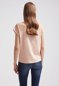 Světle růžové tričko s krátkým rukávem a průhledným puntíkovaným zadním panelem. Hladký materiál a volný střih, kombinováno s tmavě modrými džíny.