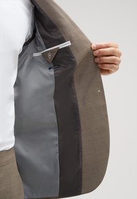 Osoba trzymająca otwartą brązową marynarkę, ukazującą szaro-czarne wnętrze z metką "Extra Slim Fit".