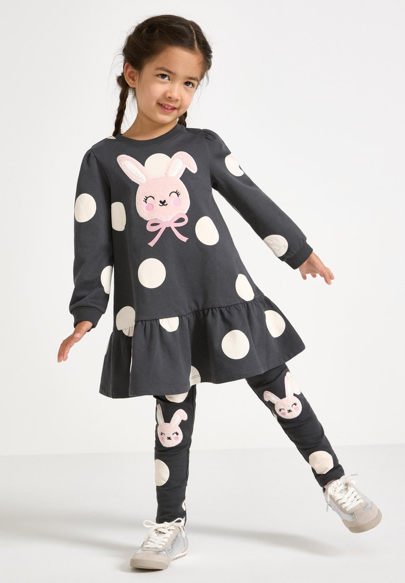 Grijze katoenen jurk met grote witte stippen, voorzien van een konijn-applicatie en een gerimpelde zoom. Gecombineerd met leggings versierd met konijnengezichten.