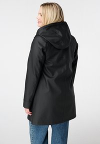 Zwarte waterdichte jas met een capuchon, zachte textuur, iets getailleerde pasvorm en naaddetail op de rug, gecombineerd met lichtblauwe spijkerbroek.