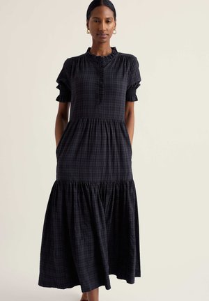 Žena v dlhých čiernych károvaných šatách s krátkymi nafúknutými rukávmi, volánikovým golierom, gombíkmi na živôtiku a nariasenou sukňou v úrovniach, ruky v vreckách.