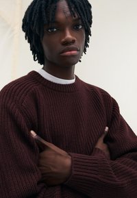 Burgundy ribbad stickad tröja med rund halsringning, med texturerad tyg och långa ärmar, bärs av en person som har armarna över kors.