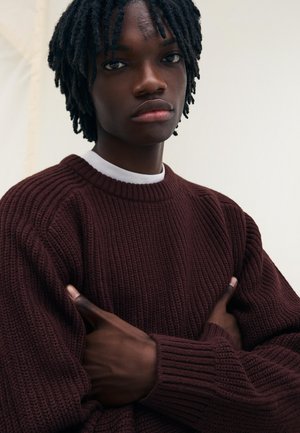 Bordowy sweter z prążkowanego dzianiny z okrągłym dekoltem, z teksturowanej tkaniny i długimi rękawami, noszony przez osobę z założonymi na siebie rękoma.