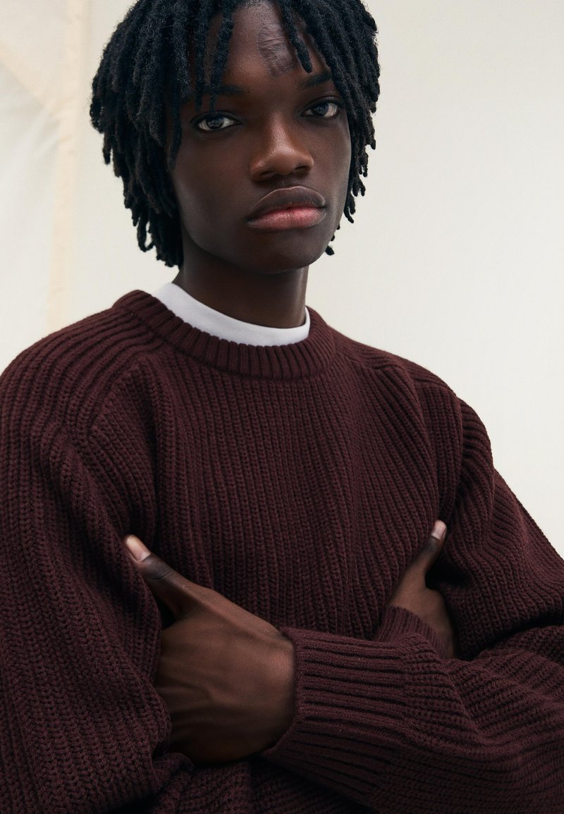 Burgundy ribbad stickad tröja med rund halsringning, med texturerad tyg och långa ärmar, bärs av en person som har armarna över kors.