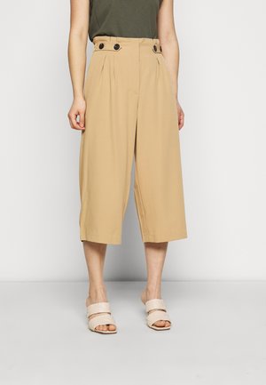 Beige wijde cropped broek met hoge taille en knopen, gedragen met een donkergroene top en beige sandalen met hak.