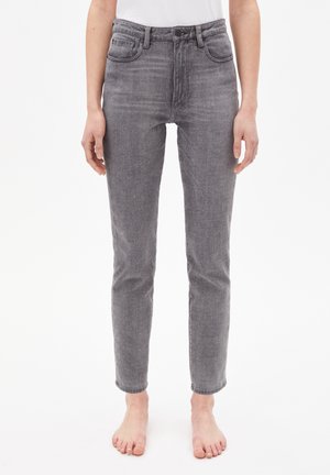 Jean en denim gris coupe droite porté pieds nus par une personne debout devant un fond blanc uni.