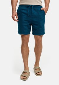 Türkise Shorts mit lockerer Passform, Kordelzugbund und seitlichen Taschen, aus strukturiertem Stoff. Kombiniert mit leichten Sandalen.