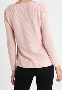 Femme portant un pull long à manches longues rose pâle à côtes et un pantalon noir, vue de dos sur un fond blanc uni.