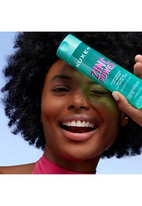 Bottiglia turchese di lozione NUXE Zinc Power, con testo rosa audace, tenuta da un modello sorridente con capelli neri e texturizzati e una maglietta rosa.