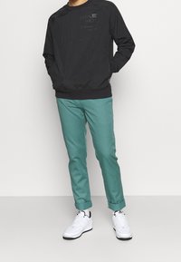 Sweatshirt noir à col rond avec un logo discret, associé à un pantalon teal clair et des baskets blanches. Tissu doux, coupe décontractée et poignets élastiques.
