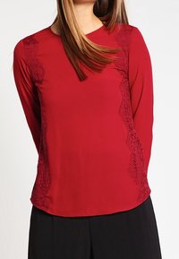 Haut rouge à manches longues avec des accents en dentelle sur les côtés, col rond, et un tissu lisse légèrement texturé. L'ourlet est arrondi.