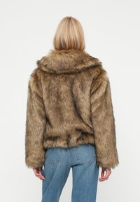 Giacca in ecopelliccia marrone con colletto largo e maniche lunghe, caratterizzata da una finitura morbida e testurizzata. Abbinata a jeans in denim azzurro chiaro.