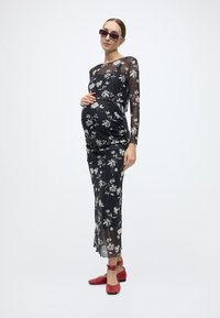 Μακρυμάνικο μαύρο φόρεμα με floral σχέδιο, εφαρμοστή σιλουέτα, διαφανές επίστρωμα και ρυθμισμένη εμπρόσθια ραφή. Συνδυάζεται με κόκκινα επίπεδα παπούτσια.