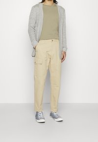 Beige Cargo-Hosen mit einem Frontknopfverschluss und seitlichen Taschen, kombiniert mit einer grauen Strickjacke und einem hellgrünen T-Shirt sowie grauen Sneakern.