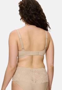 Nainen, jolla on kihara ruskea hius, pukeutuneena beigeen rintsikkasettiin ja korkeavyötäröisiin pitsialushousuihin, näkyy selkäpuolelta yksinkertaista taustaa vasten.