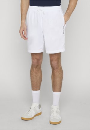 Hombre con pantalones cortos blancos con cordón y bolsillos laterales, calcetines blancos y zapatillas blancas con suelas de goma, de pie sobre un fondo liso.