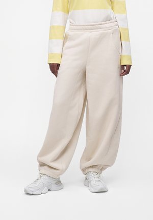 Personne portant un pantalon de jogging beige large avec des poignets élastiques, des baskets blanches, et un t-shirt à manches longues rayé jaune et blanc, debout.