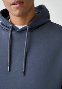 Pilkas džemperis su kapišonu, pasižymintis minkšta tekstūra, reguliuojamomis virvelėmis su metaliniais galiukais ir sluoksniuotu dizainu virš balto marškinėlio.