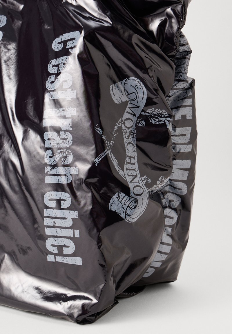 Borsa nera lucida con la scritta "C'est trash chici!" e "Moschino" stampata in grigio logorato. Presenta una texture stropicciata e accenti grafici astratti.