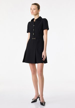 Vrouw staand, gekleed in een zwarte korte mouwen jurk met knopen, een kleine riem en zwarte puntige slingbackhakken op een witte achtergrond.