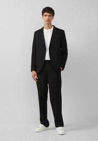 Schwarze Sakkojacke mit zwei Knöpfen und Taschen, kombiniert mit schwarzen Hosen und einem weißen T-Shirt. Getragen mit weißen Turnschuhen.