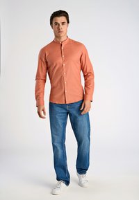 Camisa de botones de manga larga color coral con cuello mao, combinada con jeans azules y zapatillas blancas, de pie contra un fondo claro.
