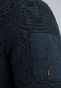 Navy trui met een gestructureerd, ribbelgebreid patroon en een soepele stoffen patch op de elleboog, met een klein geel merklabel.