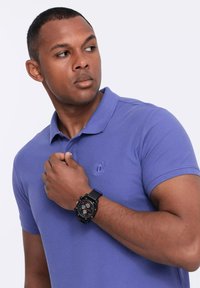Camisa polo morada con un acabado texturizado y un logo bordado, combinada con un reloj de pulsera negro que tiene una esfera de varios colores.