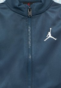 Μπλε ναυτικό μπουφάν με φερμουάρ μπροστά, απαλό ύφασμα και λευκό λογότυπο Jumpman στο δεξί μέρος. Διακριτική λάμψη σε όλο το ύφασμα.