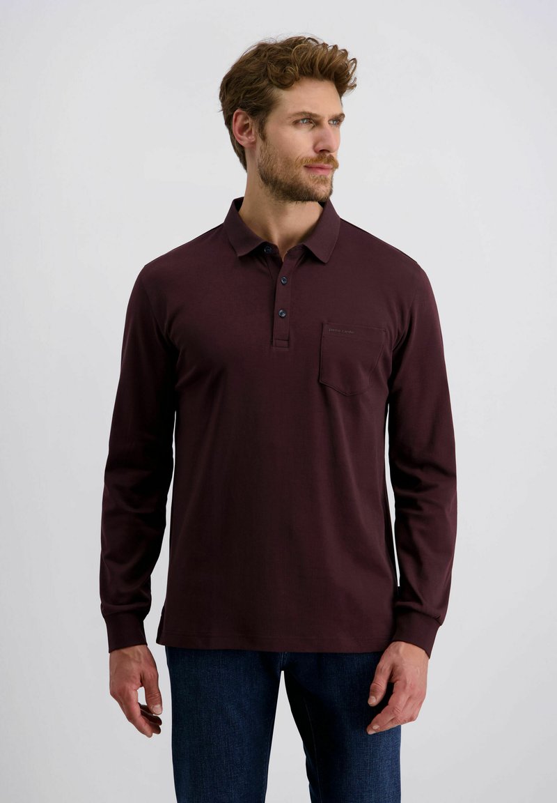 Mann mit hellbraunen Haaren, der ein marsala-farbenes Langarm-Polohemd und dunkle Jeans trägt und nach rechts blickt, vor einem schlichten Hintergrund.