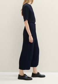 Žena v námořnickém tričku s krátkým rukávem a širokých zkrácených kalhotách, doplněná černými šněrovacími botami, stojící bokem na jednobarevném světlém pozadí.