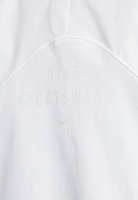 Hvit stoff med en glatt tekstur har "Nike Sportswear"-logoen i lys grå. En liten Nike swoosh er også tilstede.
