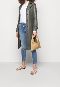 Harmaa nahkatrenssi, jossa on kaksirivinen etuosa, valkoinen napitettava paita, vaaleansiniset skinny-farkut ja tan-värinen käsilaukku, jossa on solmukahva. Valkoiset remmissandaalit.