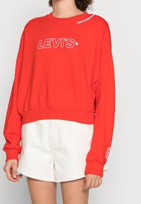 Felpa rossa con logo "LEVI'S" bianco, vestibilità ampia, scollo a girocollo e polsini a costine. Abbinata a pantaloni corti bianchi. Materiale in cotone texturizzato.