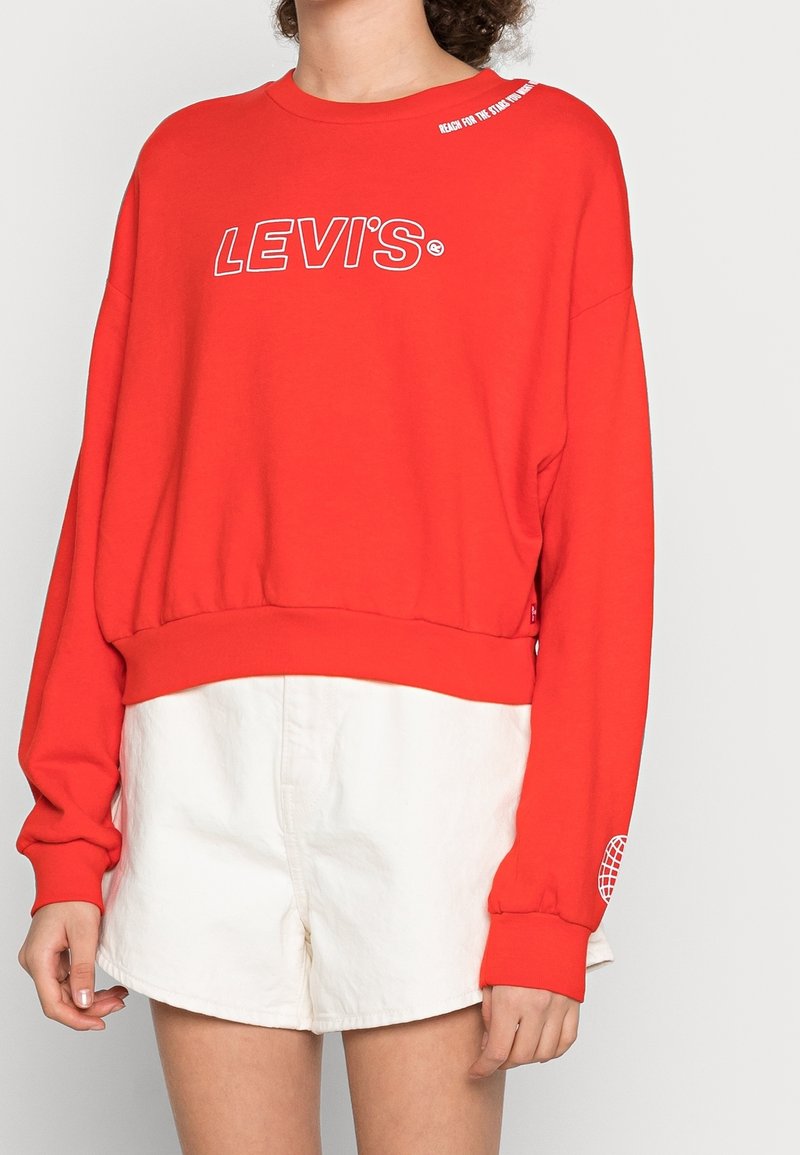 Felpa rossa con logo "LEVI'S" bianco, vestibilità ampia, scollo a girocollo e polsini a costine. Abbinata a pantaloni corti bianchi. Materiale in cotone texturizzato.