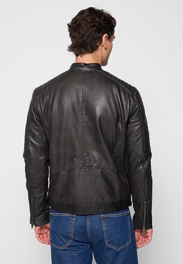 Giacca di pelle nera con spalle trapuntate e polsini con zip, caratterizzata da un design aderente e una texture liscia. Indossata con jeans blu.