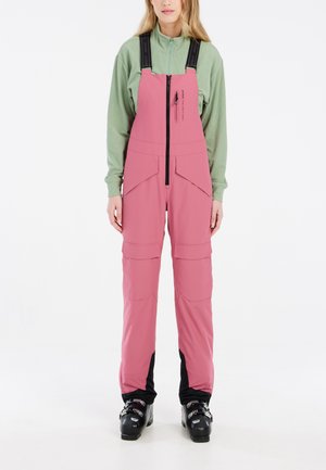 Pink ski overalls with adjustable straps, zippered front, multiple pockets, and black accents at the ankles. Worn with black ski boots.