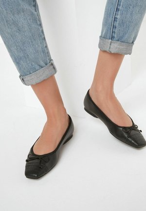Pieds portant des chaussures plates noires avec détail nœud et bout texturé, associées à un jean bleu clair retroussé sur fond blanc.