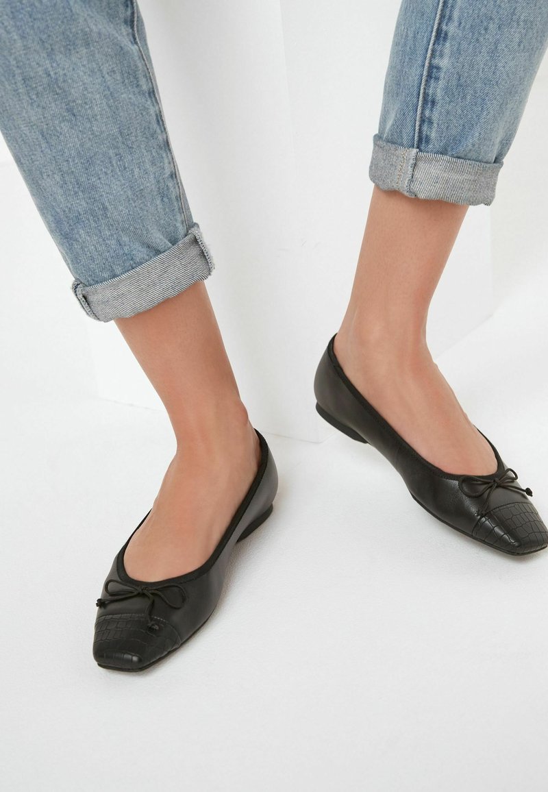 Pieds portant des chaussures plates noires avec détail nœud et bout texturé, associées à un jean bleu clair retroussé sur fond blanc.