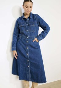 Tumši zila džinsu kleita ar garām piedurknēm, pogu aizdari un divām krūšu kabatām. Audums ir gluds un plūstošs siluets.