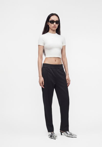 Γυναίκα με μαύρα γυαλιά ηλίου, λευκό cropped t-shirt, μαύρο παντελόνι και ασημί μυτερές ψηλοτάκουνες γόβες, να στέκεται μπροστά σε ένα μονόχρωμο φόντο.