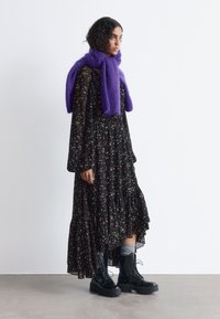 Schwarzes Blumenkleid mit langen Ärmeln und gestuftem Saum, ergänzt durch einen dicken lila Schal und klobige schwarze Schnürstiefel.